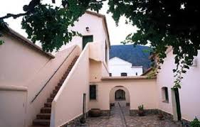 Courtyard of a light-colored building with stairs, stone pavement, potted plants, and leafy trees.