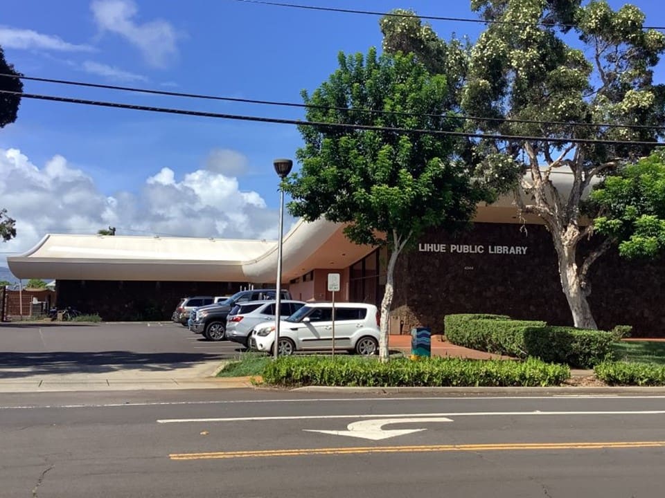 Lihue Public Library building with curved roof, greenery, parking area, and clear skies with clouds.