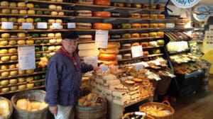 Man standing in a cheese shop surrounded by various cheeses displayed on shelves and in baskets