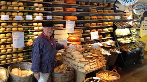 Man standing in a cheese shop surrounded by various cheeses displayed on shelves and in baskets
