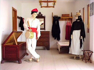 Room with two mannequins in traditional attire, wooden furniture, open chest, dresser, and coatrack.