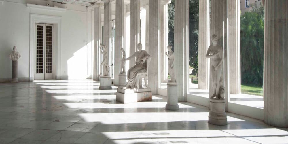 Marble statues in a sunlit hallway with arched windows and polished stone flooring.