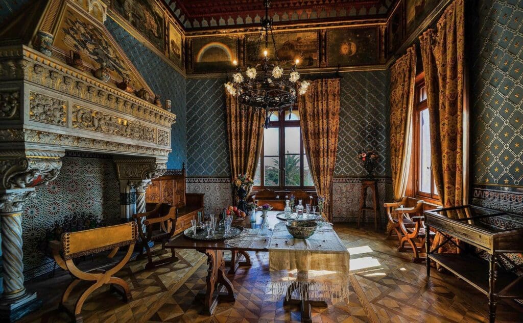 Ornately decorated room with stone fireplace, medieval furniture, chandelier, and patterned wallpaper.