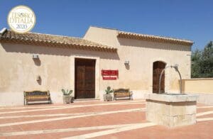 Mediterranean-style building with wooden doors, benches, potted plants, stone well, and tiled roof.