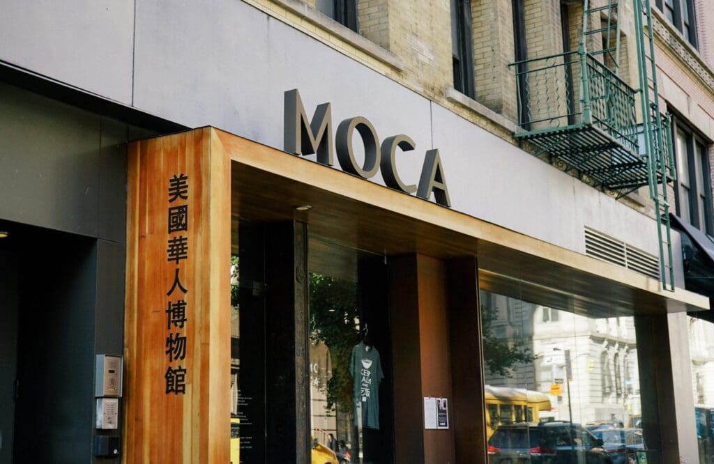 Museum entrance in an urban area with "MOCA" sign and Chinese characters on a wooden panel.