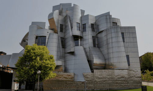 Modern abstract building with curved metallic surfaces, windows, and greenery including a tree.