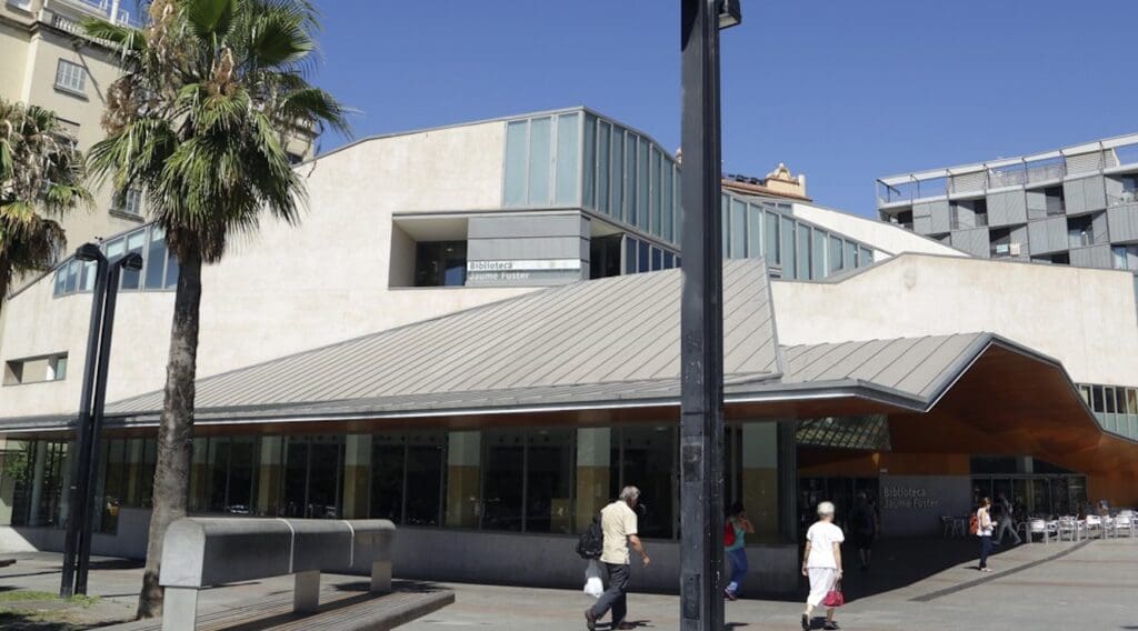 Modern building with angular design, large windows, palm trees, pedestrians, and "Biblioteca Jaume Fuster" sign.