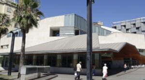 Modern building with angular design, large windows, palm trees, pedestrians, and "Biblioteca Jaume Fuster" sign.