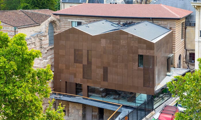 Modern angular building with semi-transparent and solid brown panels beside traditional stone structure.