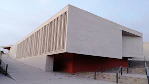 Modern, angular building with vertical slats, elevated with stairs, red section near base, clear sky.
