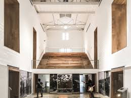 Modern art gallery with high ceiling, large windows, wooden staircase, and visitors viewing exhibits.