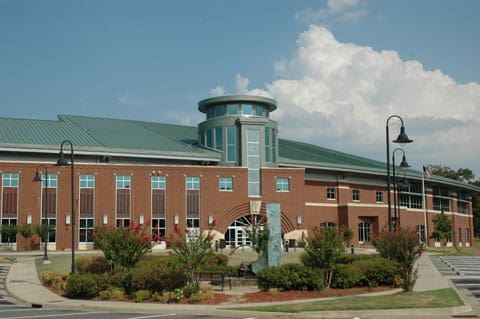 Modern brick building with green roof, central cylindrical section, landscaped area, shrubbery, lampposts, sculpture.