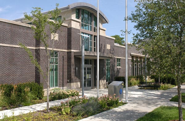 Modern brick building with large windows, central entrance, landscaped front, trees, and flagpoles.