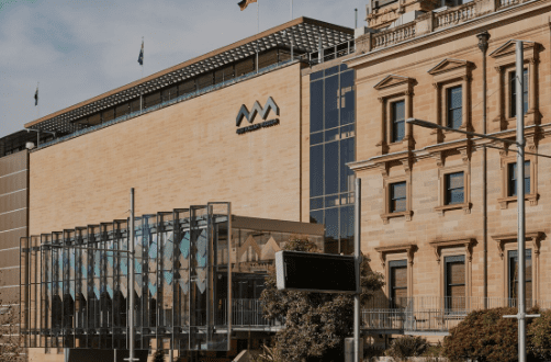Modern yellowish-brown building with glass entryway, next to older classical structure with flags.