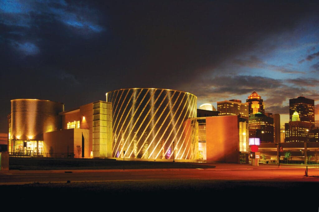 Modern building with cylindrical elements and striped lighting against a dark urban cityscape sky.