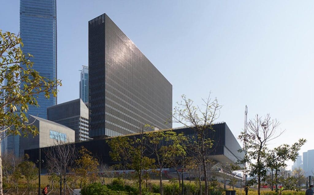 Modern building with dark facade, tall urban structures in background, and sparse trees in foreground.