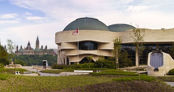 Modern building with rounded dome roof, greenery, Canadian flag, and Gothic-style structures in background.