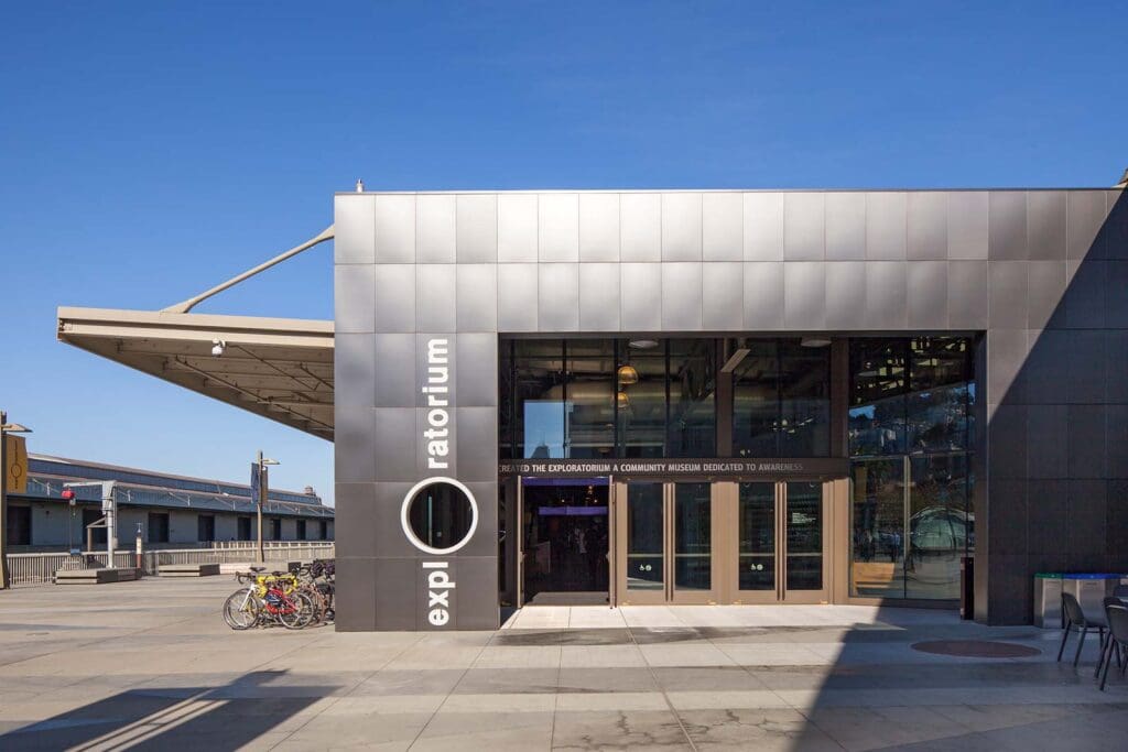 Entrance of modern building with metallic facade, glass doors, "exploratorium" sign, and bike racks
