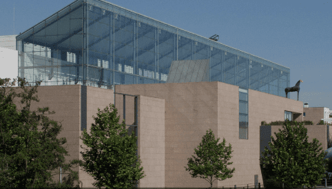 Modern building with glass upper level, beige stone walls, trees, and black horse statue on top.