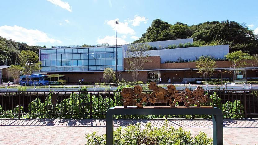 Modern building with glass windows in front of a hillside, a sculpture, and a blue and yellow bus.
