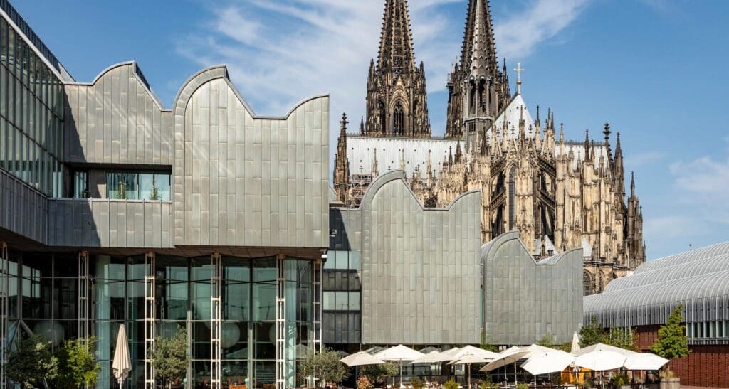 Modern building with outdoor seating in front of Gothic cathedral with ornate spires against blue sky.