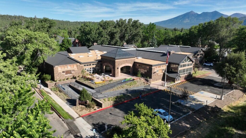 Modern stone and metal building with outdoor seating, parking, and pathways, surrounded by greenery and mountains.