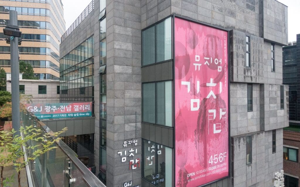 Modern multi-story building with Kimchikan Museum signage, large pink banner, G&J Gwangju-Jeonnam Gallery.