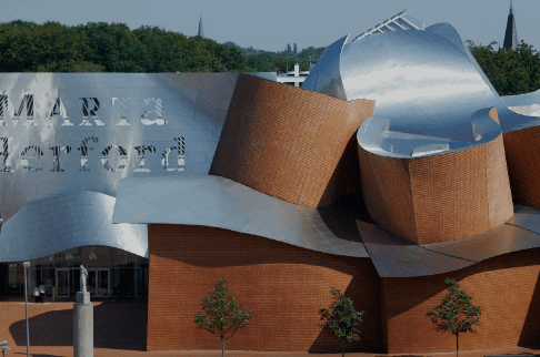 Modern building with unique metallic and brick structures, trees in front, greenery, and distant spires.
