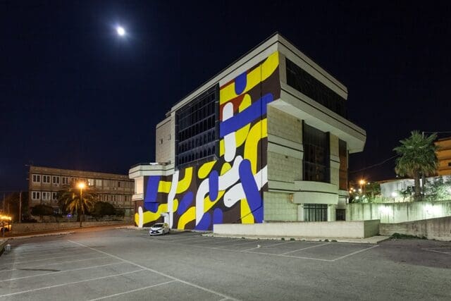 Modern multi-story building with a vibrant mural, illuminated at night, in an empty parking area.