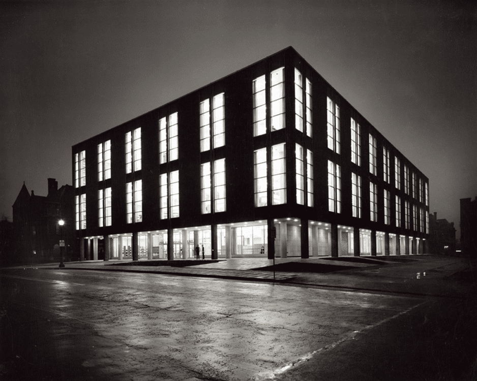 Modern four-story building with large illuminated windows on a wet urban street corner at night.