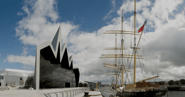 Modern angular building next to an old sailing ship at a waterfront under a partially cloudy sky.