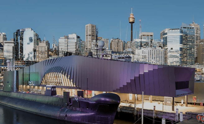 Modern building with purple, geometric facade, submarine docked at urban waterfront, skyscrapers behind