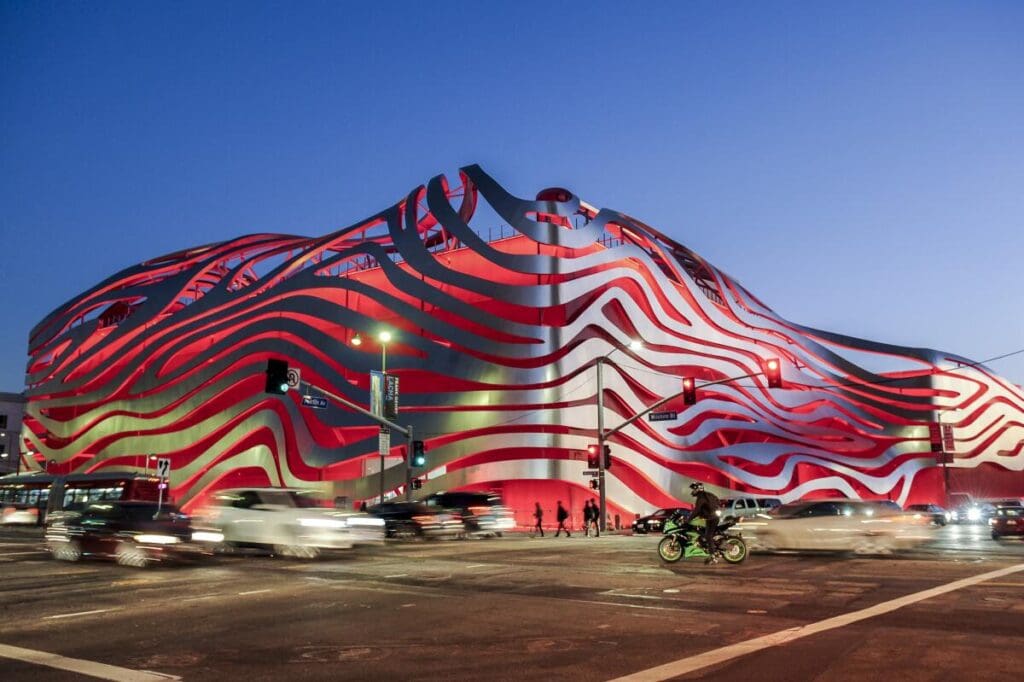 A modern building with red and white wave-like facade at a busy urban intersection with traffic.