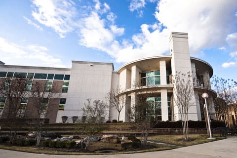 Modern multi-story building with stone and glass facade, circular entrance, trees, and a paved walkway.