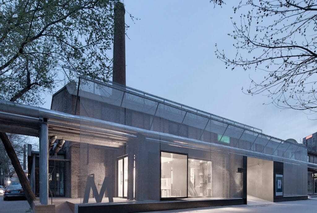 Modern building with a transparent mesh facade, trees, and industrial elements, next to a chimney.