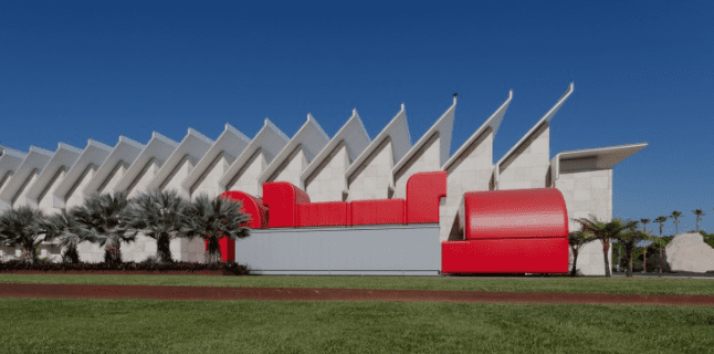 Modern building with zigzag roofline and red accents on a grassy area with palm trees under blue sky
