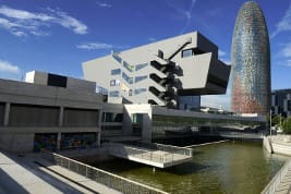Modern buildings with unique designs beside a reflective waterbody under a clear blue sky.