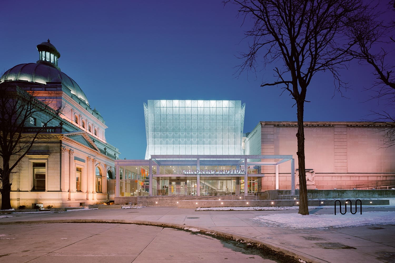 Modern glass-fronted children's museum and classical domed building lit up at night with snow.
