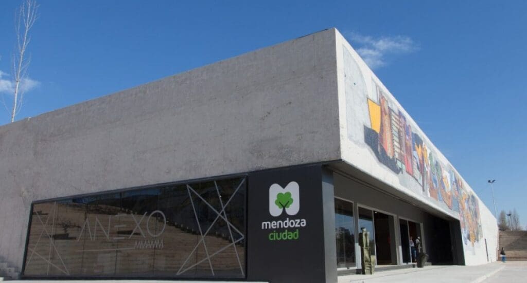 Modern concrete building with flat roof, large glass window, colorful mural, and "mendoza ciudad" sign.
