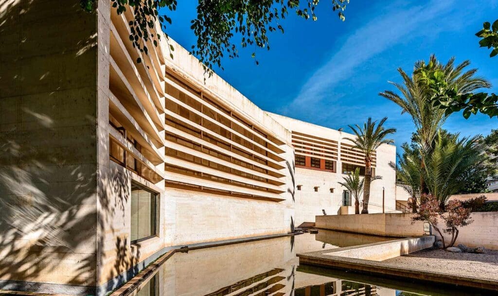 Modern concrete building with horizontal slats, palm trees, and water feature under clear blue sky.