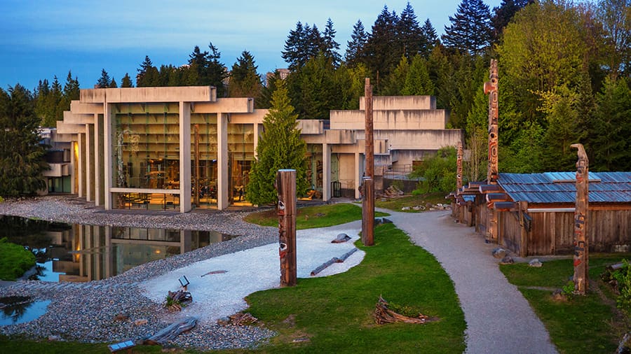 Modern concrete building with large windows, pond, gravel pathway, totem poles, and wooden cabins.