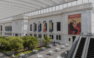 Spacious modern courtyard with high glass ceiling, tables, plants, banners on wall, and an escalator.