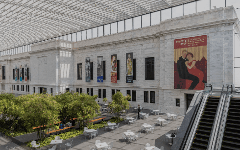 Spacious modern courtyard with high glass ceiling, tables, plants, banners on wall, and an escalator.