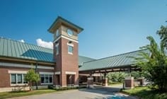 Modern building with green metal roof, brick and glass exterior, clock tower, canopy entrance, trees.