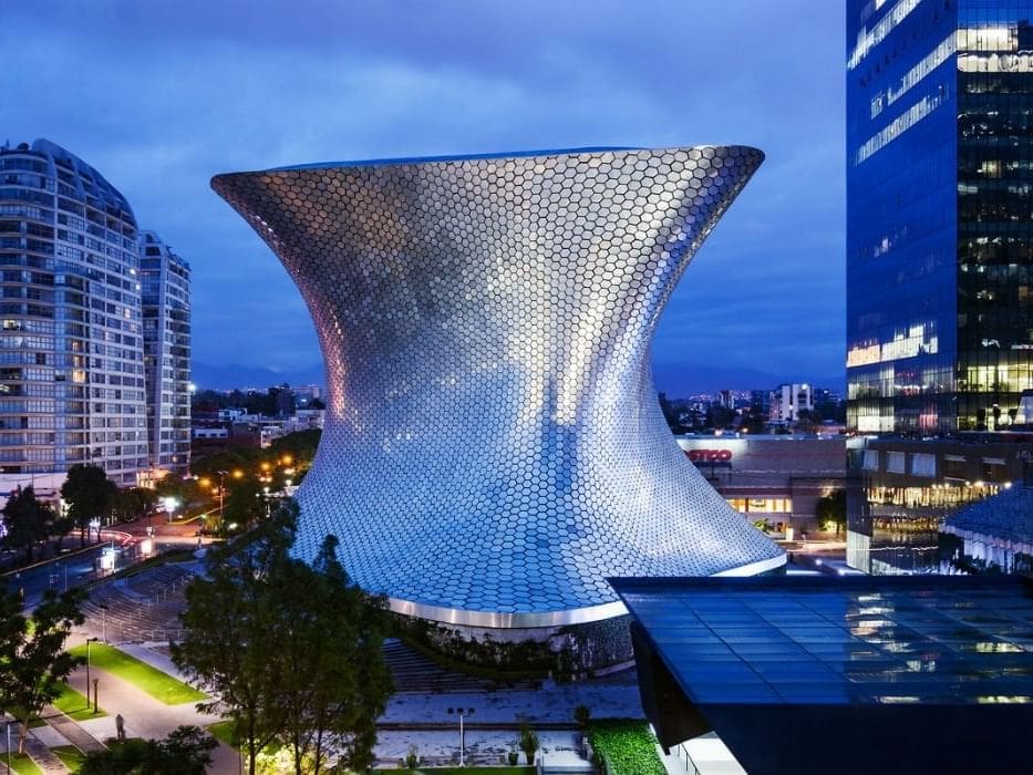 A modern, hourglass-shaped building with a hexagonal pattern illuminated in an evening urban setting.