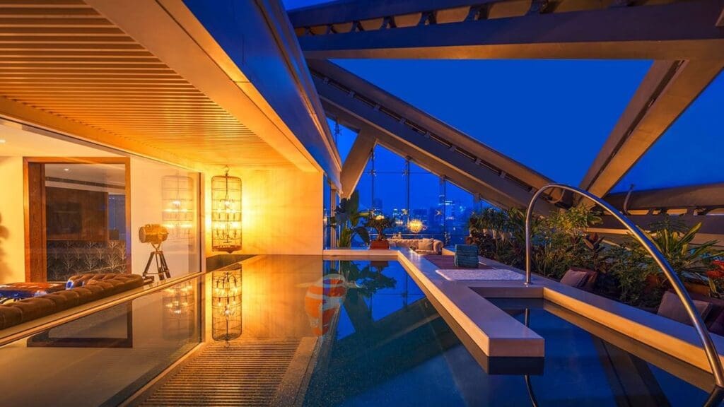 Modern indoor area with pool, seating, geometric architecture, warm lights, greenery, and city view.