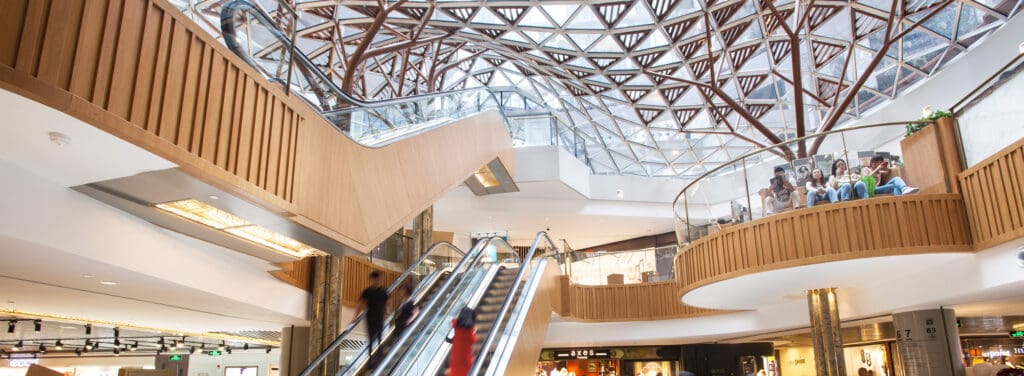 Modern indoor shopping mall with open layout, escalators, geometric glass ceiling, and seating areas.