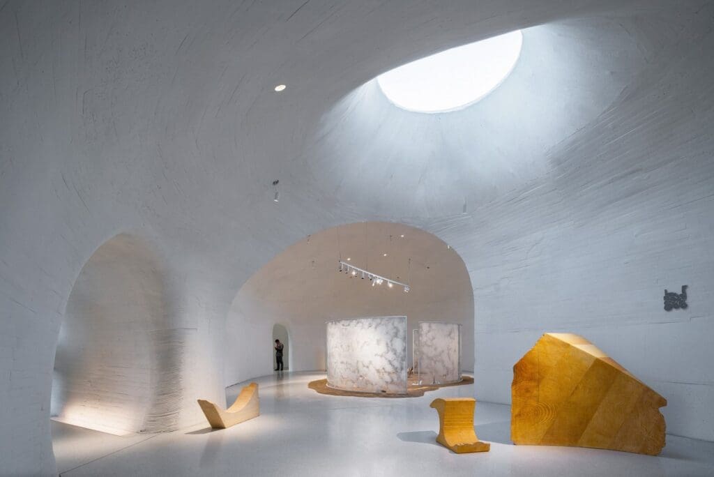 Modern interior with curved white walls, skylight, organic wood and marble structures, figure in background.