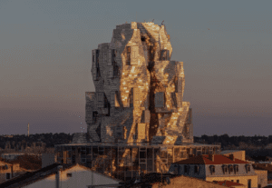 Modern, irregularly designed building with metallic panels and glass against sunset, amid rooftops and trees.