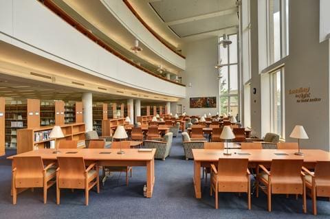 Modern library interior with wooden desks, study lamps, large windows, and bookshelves lining walls.